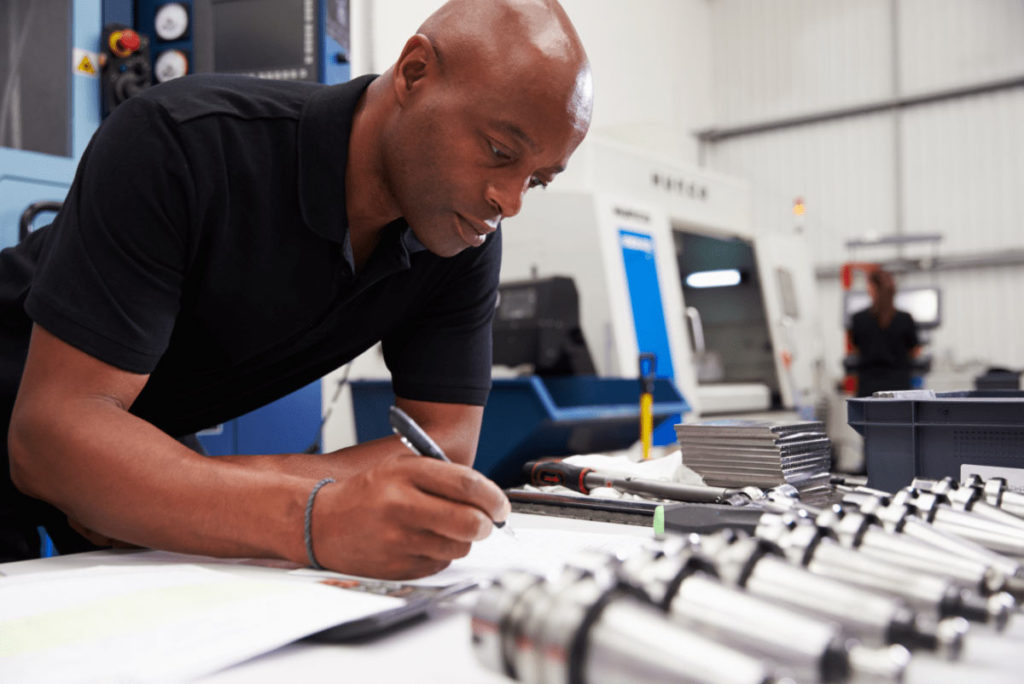 This picture shows a man in a black polo looking down at some engineering parts while writing in his notebook.