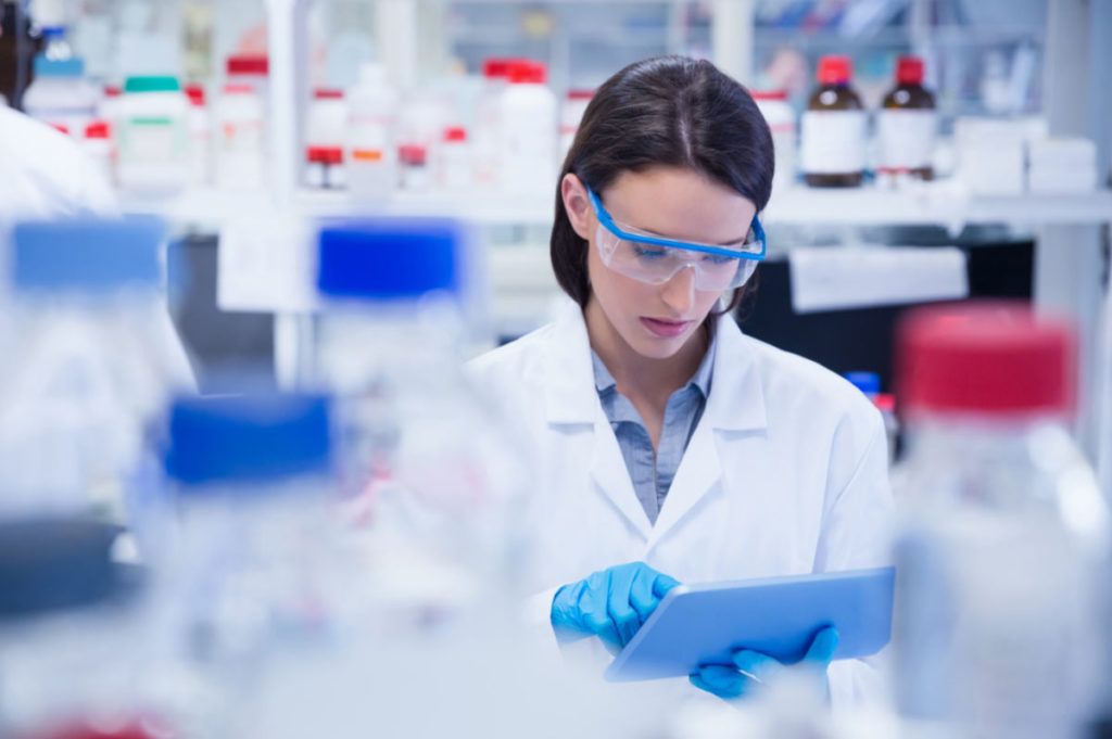 This picture shows a woman in lab coat, standing in a lab, looking down and touching her tablet.
